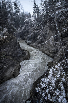 View of a turbulent river carves its path through a steep, rocky gorge, framed by snow-dusted trees under a muted sky, Bavaria, Germany.