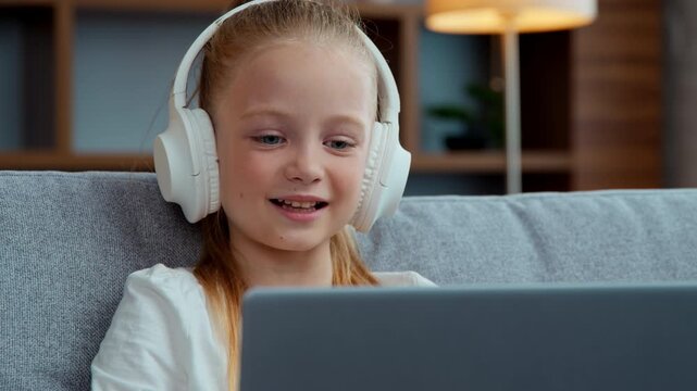 Caucasian smiling little child girl in headphones looking laptop at home happy kid daughter e-learning studying online watch lesson listen audio music playing computer game watching movie cartoons