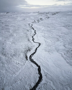 Aerial view of a dark river snaking through a vast, snow-covered landscape under a muted sky, creating a stark contrast between the dark water and the white expanse., Vik, Western Region, Iceland.