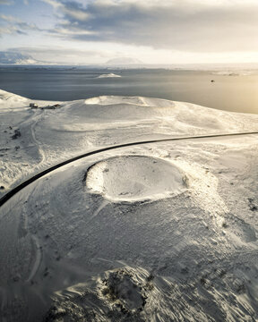 Aerial view of a stark, snow-covered landscape punctuated by volcanic cones, with a dark ribbon of road cutting through, leading towards the distant ocean, Vik, Western Region, Iceland.