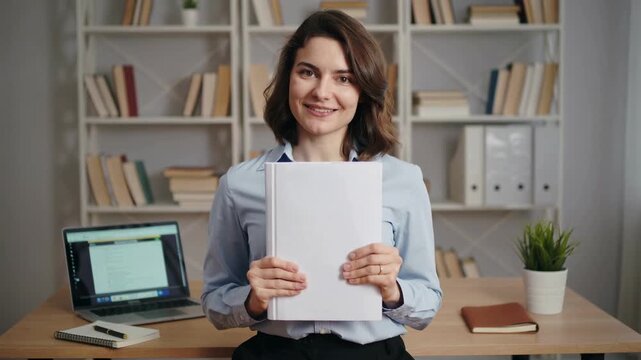 A young woman smiling and holding a stack of papers while seated at a desk, with a computer and bookshelf in the background