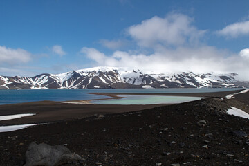 View of dark volcanic sands meet turquoise waters beneath snow-capped mountains under a sky brushed with wisps of clouds, Deception Island, Antarctica.