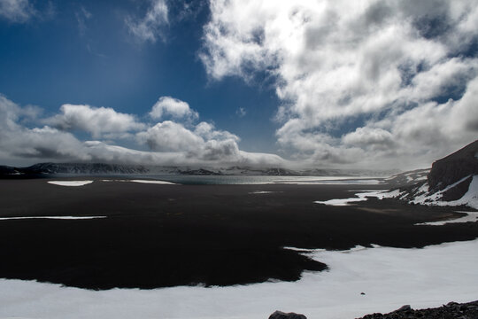 View of dark volcanic sands meeting icy waters under a sky swirling with dramatic clouds, creating a stark contrast of elements, Deception Island, Antarctica.