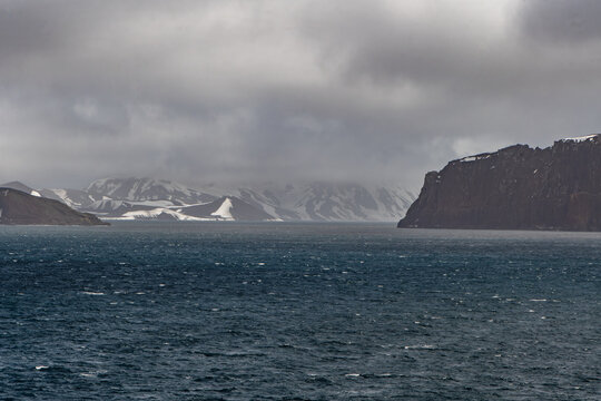 View of dark, choppy waters meet rugged, snow-dusted volcanic shores under a brooding sky, a stark contrast of textures and tones that defines a remote, icy landscape, Deception Island, Antarctica.