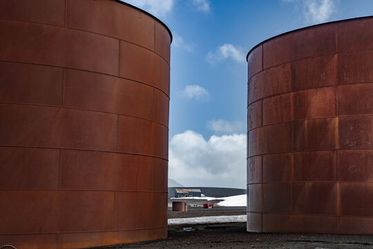 View of colossal, weathered rust-colored tanks stand sentinel against a backdrop of stark, volcanic earth and a sky streaked with wisps of clouds, Deception Island, Antarctica.