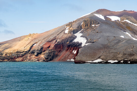 View of rugged, volcanic slopes descend into the frigid waters, a stark contrast of fiery red, gray rock, and icy patches, Deception Island, Antarctica.