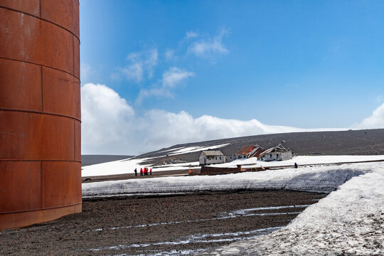 View of rust-colored tanks juxtapose against desolate volcanic sands and remnants of snow, with distant buildings under a bright sky, Deception Island, Antarctica.