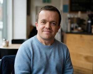 A man is sitting inside a cafe. He has a friendly smile and is wearing a blue sweater. The cafe has wooden furniture and a casual atmosphere.