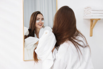 Beautiful woman in bathrobe with eye patches near mirror in bathroom