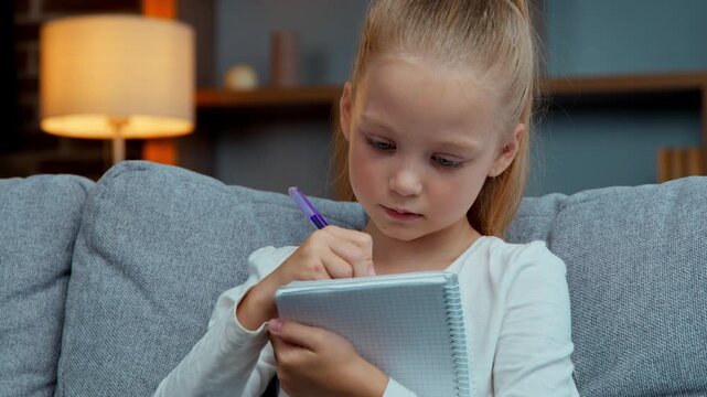 Caucasian focused little kid daughter girl child writing homework at home elementary primary learning studying preparing school exams write in notebook copybook cute pupil schoolgirl education study