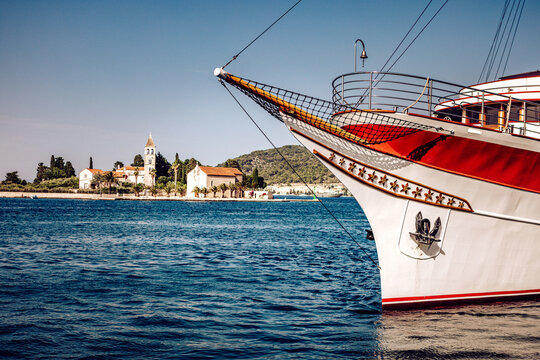 View of the bow of a classic ship with red accents meets the tranquil, shimmering sea, with a distant island and buildings, Vis, Split-Dalmatia County, Croatia.