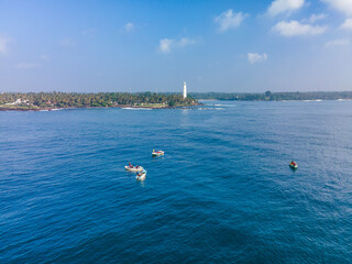 Aerial drone photo of Dondra Head Lighthouse in Sri Lanka with traditional fishing boats and fishermen in the foreground. Tall white lighthouse standing on the southernmost point of the island.