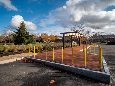 View of a wooden deck with yellow railings under a pergola, contrasting with a green lawn and scattered trees under a cloudy sky, Apollo Park, Oregon, United States.