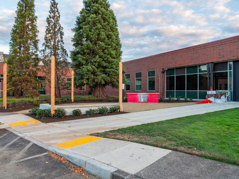 View of red brick building with large windows, lush green grass, trees, and a concrete sidewalk, contrasting with the blue sky, Apollo Park, Oregon, United States.