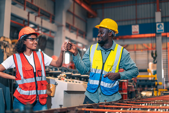 African factory worker man woman fist bump team success. Industrial engineer partner male female knuckle bump power in manufacturing plant. Safety helmet vest heavy metal machine background. Diversity