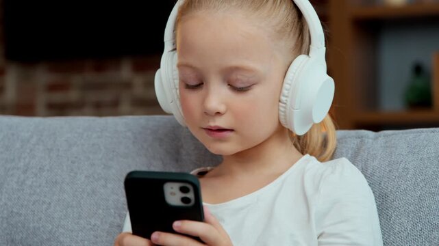 Caucasian little girl child kid listening music in headphones using mobile phone app playing virtual game daughter adorable baby in earphones look at smartphone dancing moving enjoy song listen audio