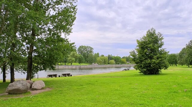 The river flows through the city among grassy lawns with aspens, willows and lime trees growing on them. There are boulders. Fences, benches and lampposts are installed on the concreted shores. Cloudy