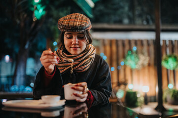 A young woman in a plaid hat and striped scarf lifts a spoon toward her tea. She sits at a reflective outdoor cafe at night.