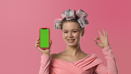 A woman in a pink blouse, with curlers shaped like banknotes in her hair, stands against a pink background, holding a smartphone with a green screen and making an OK gesture.