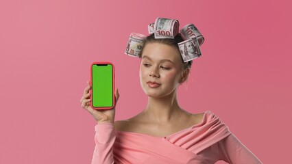 A woman in a pink blouse, with curlers in the shape of banknotes in her hair against a pink background, holds a smartphone with a green screen. A humorous concept of wealth.