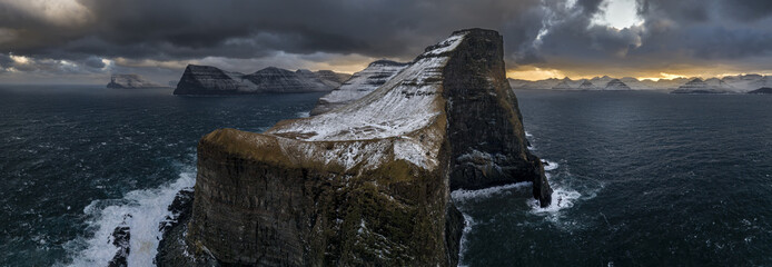 Aerial view of dramatic cliffs rise from the turbulent sea, their snow-dusted peaks catching the fading light under a brooding sky, Vagar, Vagar, Faroe Islands.