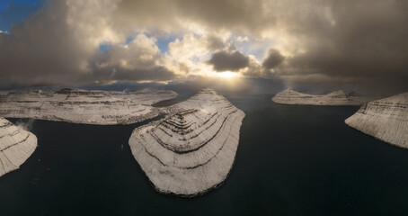 Aerial view of snow-dusted, layered islands rise majestically from the dark waters under a dramatic sky, Vagar, Vagar, Faroe Islands.