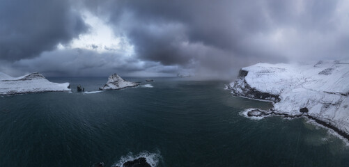 Aerial view of stark, snow-laden cliffs meet the turbulent, dark sea under a brooding sky, creating a dramatic contrast of light and shadow, Vagar, Vagar, Faroe Islands.