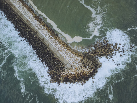 Aerial view of the breakwater where the dark, weathered rocks meet the restless, foamy sea, creating a mesmerizing dance of power and serenity, Ericeira, Lisbon, Portugal.