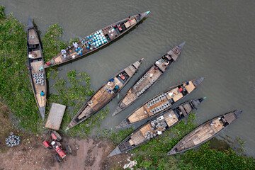 Sirajganj, Bangladesh - 12 July 2020: Aerial view of traditional wooden boats packed with goods, resting along the muddy riverbank, a rustic tractor nearby.