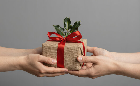 Manos entregando un regalo de Navidad envuelto en tela de yute con lazo rojo y acebo