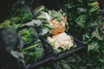 Cauliflower and leafy greens are arranged on shelves in a market. The vegetables are fresh and vibrant as people go about their shopping during the busy day