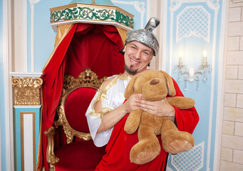 Funny smiling man weared king costume holds a teddy bear shows parody contrast