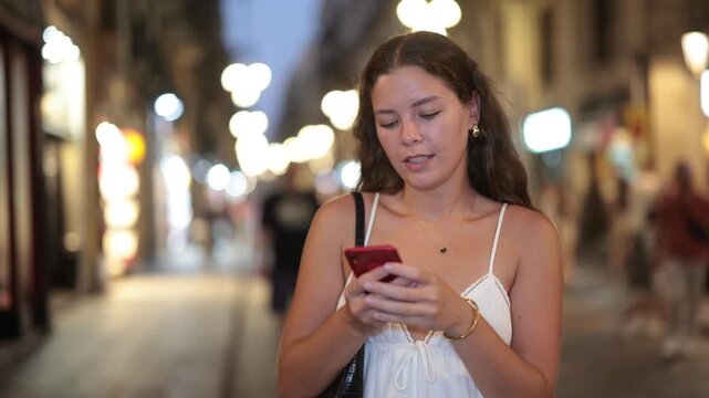 Young woman with a mobile phone in hands walks down an evening street in Barcelona, typing messages and looking at restaurant addresses in the app. Nightlife and parties in summer. High quality 4k