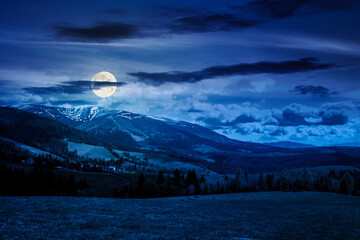 mountainous countryside landscape in spring at night. grassy meadow on top of a hill in full moon light. mountain ridge with snow capped tops. clouds on sky. finance investment or abstract concepts