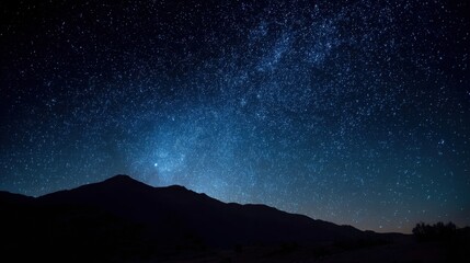A mountain range stands against the night sky filled with stars. The sky shows a rich collection of stars and a faint glow in the distance. The setting is dark and vast.