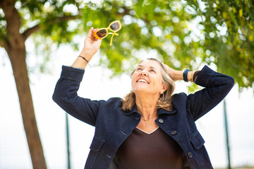 Happy woman raising sunglasses while smiling under tree