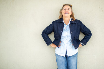 Cheerful mature woman in denim jacket laughing against neutral wall