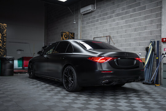 Matte black Mercedes Benz S Class sedan sits indoors, rear three quarter view, LED taillights lit, gray cinderblock walls, check floor tiles, tools and hoses visible.