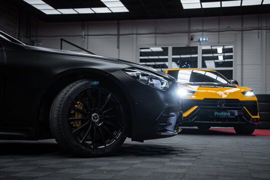 Indoor garage scene shows Mercedes S Class left and Lamborghini Urus Performante rear, headlight flares, Profilm banner, low angle, moody lighting, tiled floor, industrial lights.