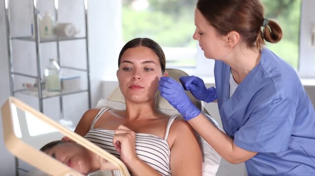 Woman client of cosmetology clinic sits in chair near doctor, looks at reflection in mirror and tells specialist about problems of contour face and in area of cheekbones