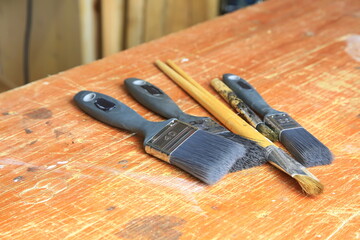 Gebrauchte Malpinsel auf zerkratztem Holzbrett: drei breite Synthetikpinsel und zwei feine Naturhaarpinsel mit Farb Sprenkeln und sichtbarer Abnutzung, Atelier-Atmosph&auml;re.