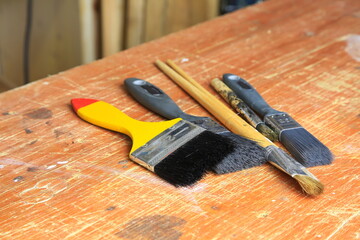Gebrauchte Malpinsel auf zerkratztem Holzbrett: drei breite Synthetikpinsel und zwei feine Naturhaarpinsel mit Farb Sprenkeln und sichtbarer Abnutzung, Atelier-Atmosph&auml;re.