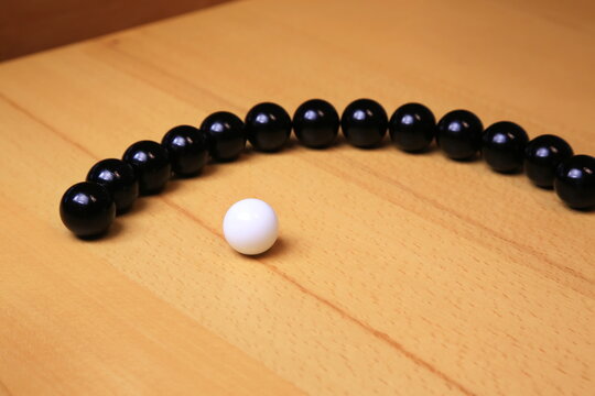 Eine wei&szlig;e Kugel steht getrennt vor einer geschwungenen Reihe schwarzer Kugeln auf Holz; der starke Hell-Dunkel-Kontrast und die reduzierte Komposition betonen Individualit&auml;t.