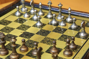 Metallisches Schachspiel auf gelb-schwarzem Brett, stilvoll arrangiert auf einem Holztisch. Silberne und bronzene Figuren in Startposition &ndash; ein Hauch von Eleganz und Strategie.