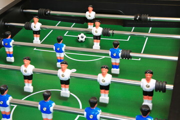 Kicker-Nahaufnahme: Ball liegt zentral auf dem gr&uuml;nen Spielfeld, flankiert von blauen und wei&szlig;en Kunststoffspielern auf Metallstangen; ein dynamischer Moment im Tischfu&szlig;ballspiel.