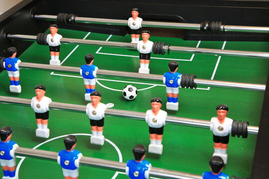 Kicker-Nahaufnahme: Ball liegt zentral auf dem gr&uuml;nen Spielfeld, flankiert von blauen und wei&szlig;en Kunststoffspielern auf Metallstangen; ein dynamischer Moment im Tischfu&szlig;ballspiel.