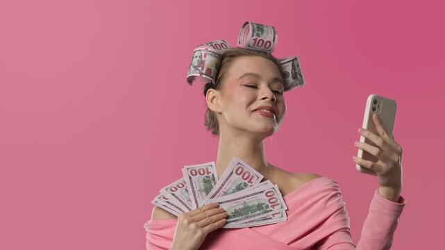 A woman in a pink blouse, with curlers shaped like banknotes in her hair, stands against a pink background, holding money in her hands and chatting happily on a video call on her smartphone.