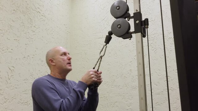 Focused bald senior man in blue sweater doing triceps cable pushdown in gym with beige textured wall background demonstrating proper form and concentration for strength training.