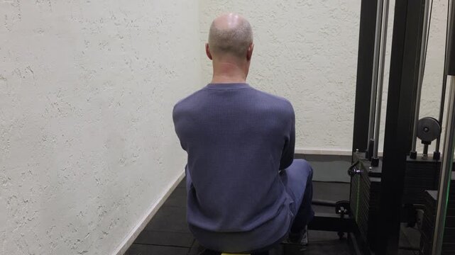 Athletic bald man in blue ribbed sweatshirt and navy pants sitting on yellow bench performing seated cable row back exercise on black multi station machine with green weight markers.
