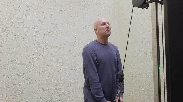 Athletic bald man in blue sweatshirt executing standing cable triceps pushdown exercise with rope attachment on black weight machine in bright modern home gym.